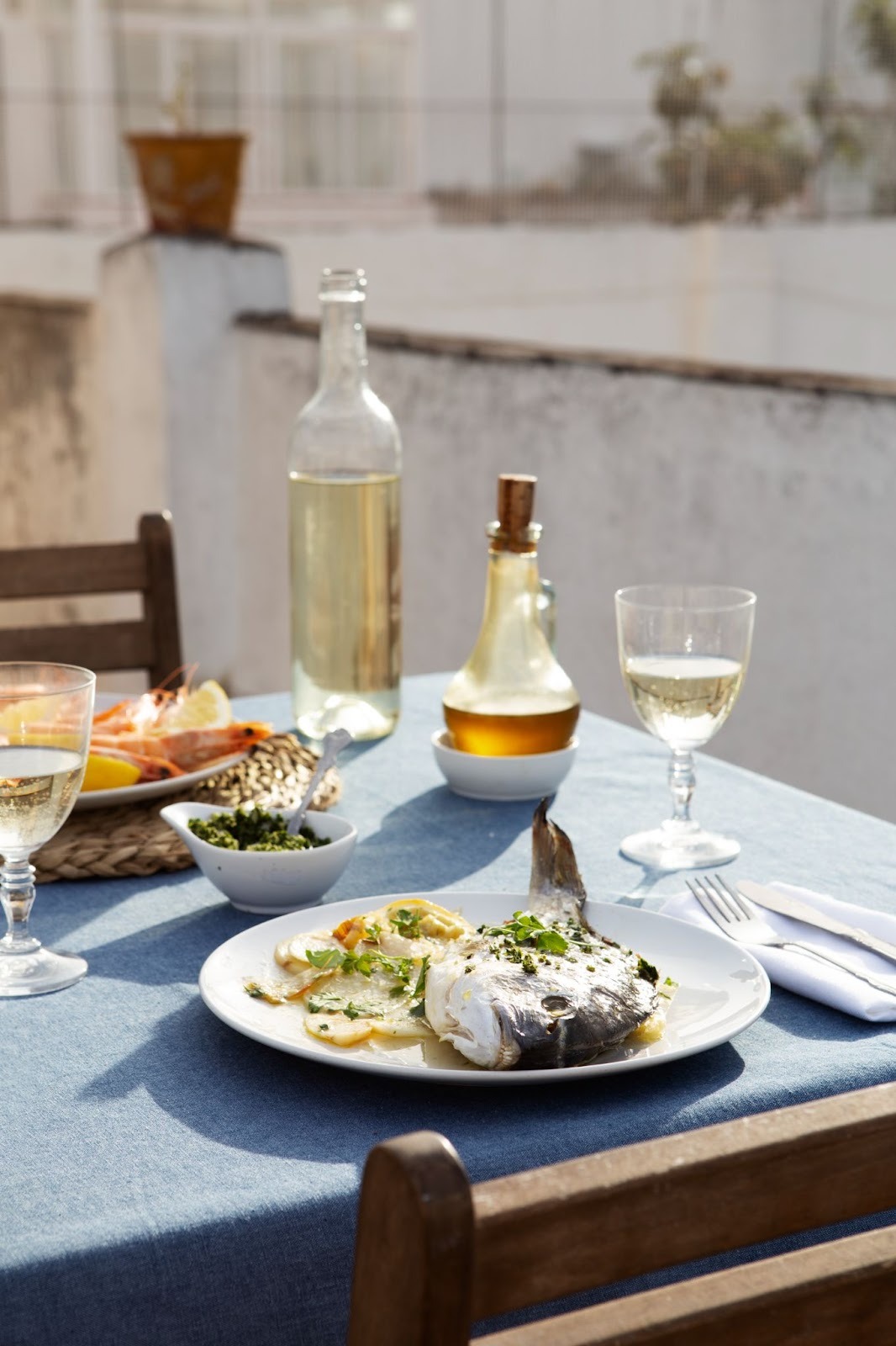 Outdoor taverna table set with a plate of cooked fish and sliced potatoes, white wine, and olive oil on a blue tablecloth.
