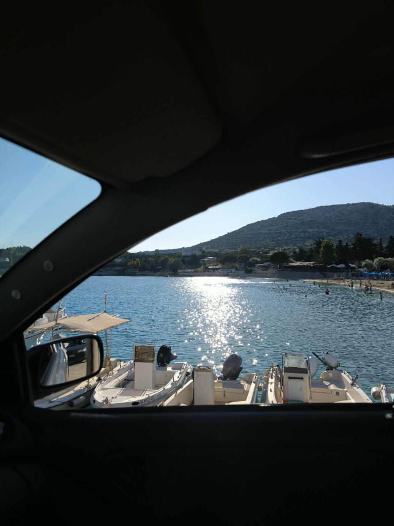 Driving in Chania and enjoying the view of Marathi Beach, from inside a car, showing small boats docked in the sparkling blue sea and green hills in the background
