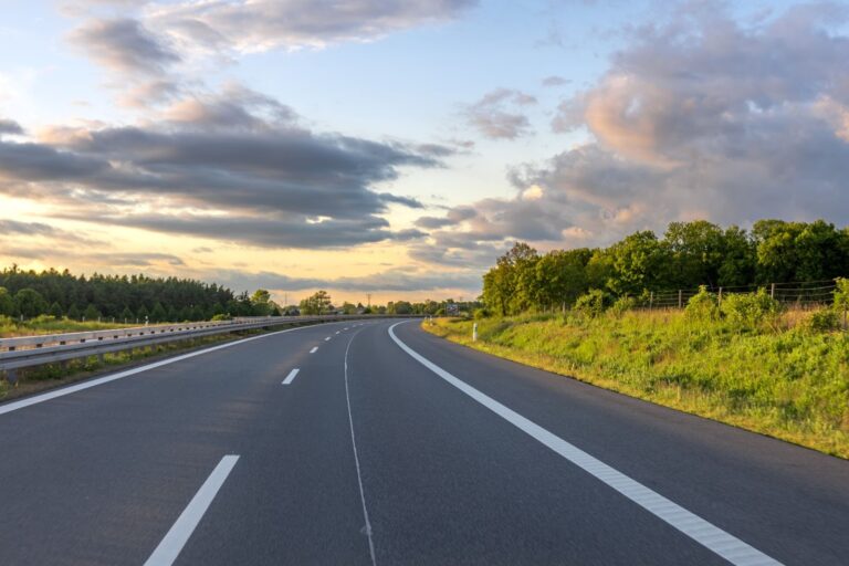 Empty two-lane highway curving through green grass and a treeline under a dramatic sunset sky with dark clouds